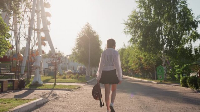 Young woman walks away from camera down sunlit street wearing headphones and carrying bag. scene features outdoor park elements like trees benches sculptures and distant amusement rides