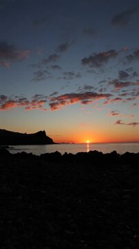 Stunning vertical sunrise over the calm Atlantic Ocean in Madeira, featuring scenic dark rocky coastline silhouettes under dramatic orange clouds at dawn. 4K vertical video.