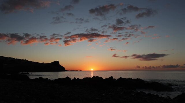 Serene sunrise over the Atlantic Ocean coast in Madeira with golden light reflecting on calm water, dramatic rocky silhouettes, and scenic clouds at dawn. 4K video.
