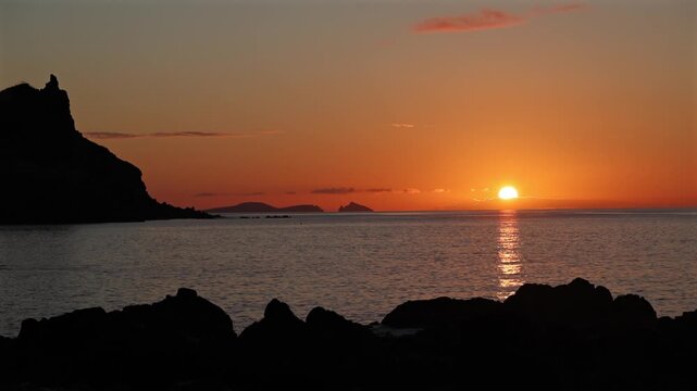 Golden glowing sunset over the calm Atlantic Ocean in Madeira with dramatic silhouetted volcanic rocks and tranquil horizon during a scenic summer dusk. 4K video.