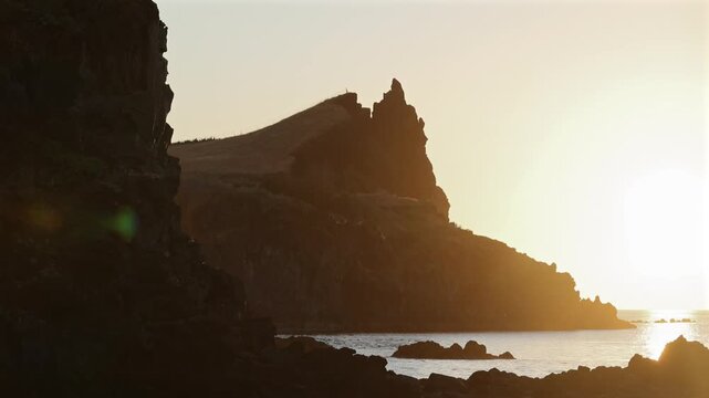 Dramatic volcanic cliffs and rugged coastline of Madeira island illuminated by warm golden hour sunlight over calm ocean waters, creating a serene mood. 4K video.