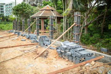 Naklejka premium Construction site with stacked concrete blocks, upright rebar columns, and timber braces beside unfinished foundation. Sand, debris, and lush trees frame small pavilion and nearby building work.