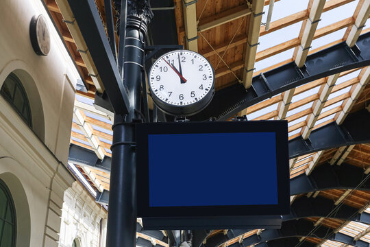 Analog station clock above blank digital information screen beneath timber and steel roof structure. Railway terminal architecture public transport infrastructure and travel background.