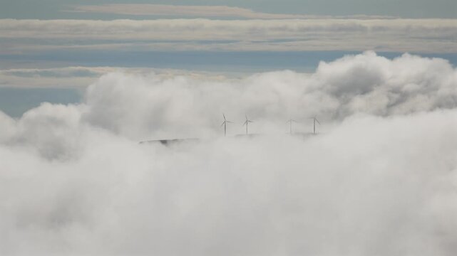 Wind turbines on high mountain plateau emerging through dense white sea of clouds in Madeira, sustainable renewable green energy concept under overcast sky. 4K video.