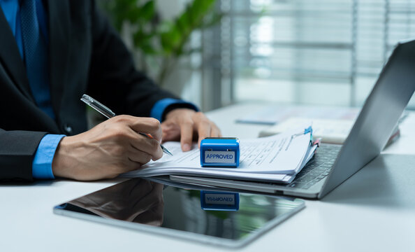 Businessman reviewing and signing approved documents beside a laptop and tablet, representing authorization, compliance, verification, corporate approval and administrative workflow.