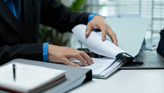 Businessman reviewing and turning pages of a detailed report beside a laptop, representing document audit, financial review, compliance, verification and corporate analysis.