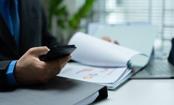 Businessman using a smartphone while reviewing reports beside a laptop, representing mobile communication, digital workflow, remote management and modern corporate productivity.