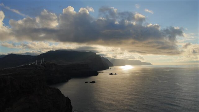 Dramatic sunrise over Madeira rugged coastline with glowing sunbeams reflecting on Atlantic ocean water, wind turbines on cliffs, and cinematic clouds. 4K video.