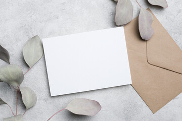 Blank white card placed on a textured gray surface beside a brown envelope and eucalyptus leaves, creating a minimalist stationery arrangement © nikavera