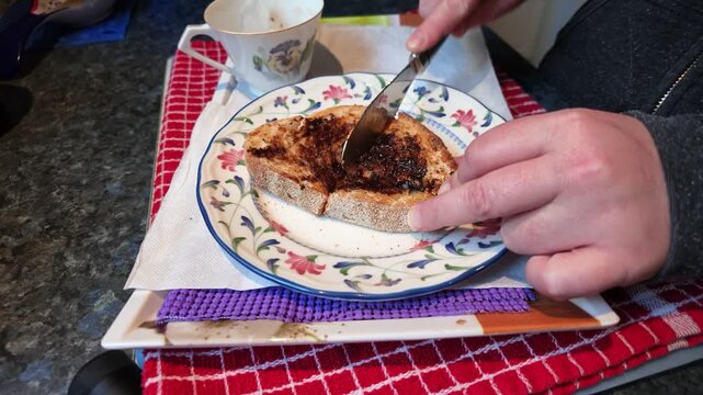 Close-up of a person's hands spreading vegemite on a slice of toasted sourdough bread and then cutting it in half, preparing breakfast