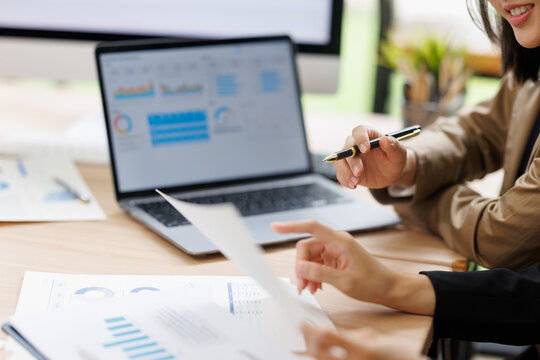 Business team analyzing financial data and market reports on laptop with charts and graphs, discussing strategy and reviewing paperwork in an office meeting
