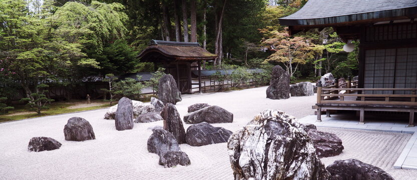日本の寺院にある静寂な枯山水庭園。白砂に描かれた波紋と配置された奇岩、背景の木々と伝統的な木造建築が調和した、静謐で精神的な禅の空間。
