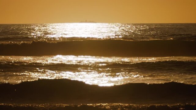 Cinematic 4K slow motion footage of gentle ocean waves rolling onto the beach during a majestic golden sunset. The warm evening light illuminates the water and sky, Guincho beach, Portugal. 