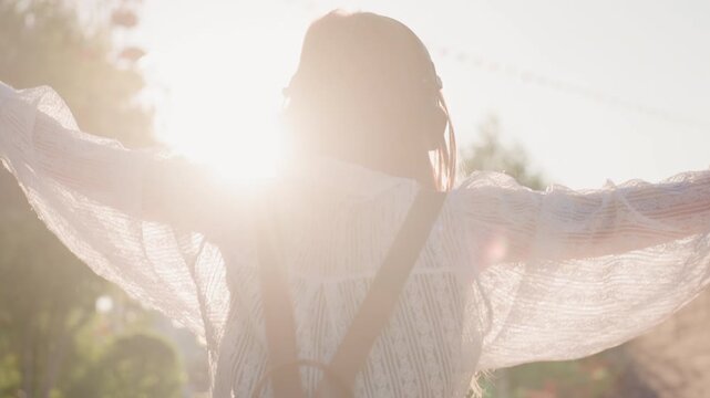 Woman with long hair wearing white lace blouse and brown suspenders raises her arms in outdoor setting during sunset sunlight creates lens flare around her silhouette. Woman wears headphones
