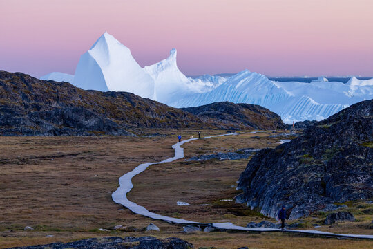 Sermermiut valley in Greenland with wooden hiking path and a giant iceberg in the background at sunset