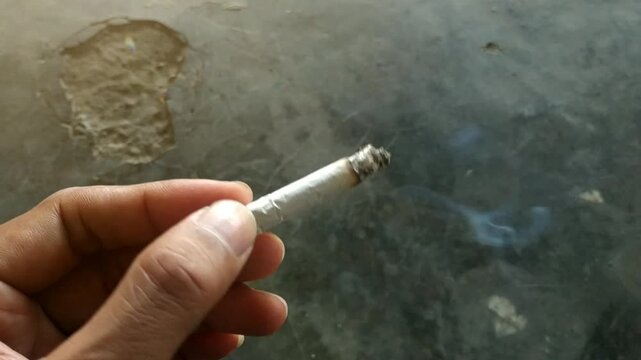 Close-up view of a person's hand holding a lit cigarette with visible ash. Represents concepts of smoking addiction, nicotine dependence, stress relief, and unhealthy lifestyle habits against a blurre