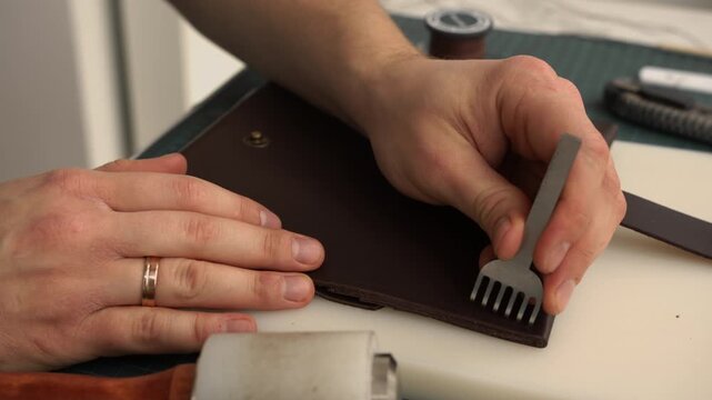 Leather craftsman hitting pricking iron with a mallet for creating stitching holes on a brown leather wallet in his workshop