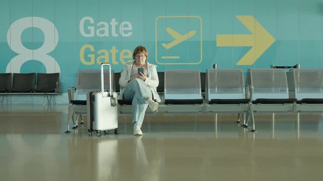 Senior woman waits for boarding at airport departure gate with carry-on bag. Mature female passenger rests and uses smartphone. Modern terminal lounge offers quiet transit waiting area.