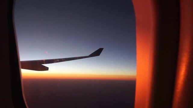 View inside passenger cabin airplane window showing wing silhouette floating over vibrant orange horizon at sunset for travel agency promo background international commercial flight footage