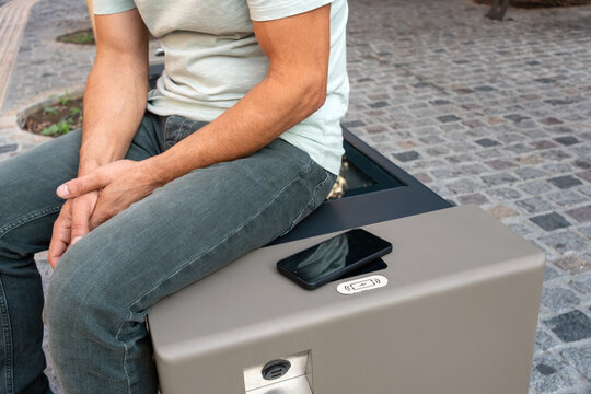 Man takes break on smart urban bench while his smartphone recharges wirelessly on solar-powered charger, combining public charging, modern technology, smart city infrastructure and everyday urban life