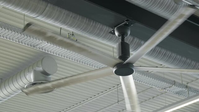 Large industrial ceiling fan rotates under high metal roof. Modern HVAC system circulates airflow for cooling. Ventilation equipment regulates indoor climate inside commercial building.