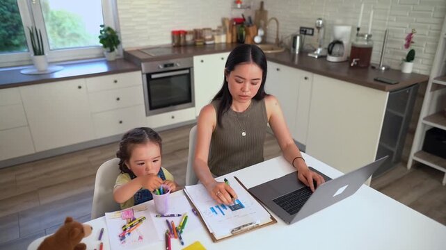 Japanese mother working from home on a laptop with her daughter drawing next to her