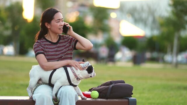 Asian woman talking on phone while petting french bulldog in park