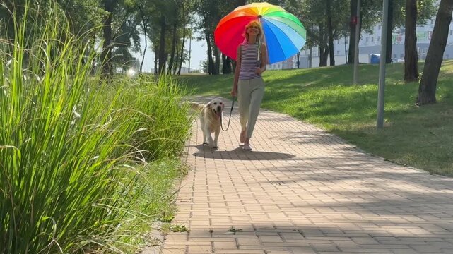 Happy woman walking Golden Retriever on leash under colorful rainbow umbrella in city park, protecting from sun, enjoying pleasant outdoor walk and bonding with her dog