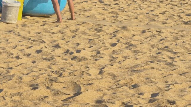 Tracking shot of a person carrying a white and a yellow bucket walking on the sandy beach toward a basket boat under morning light in Phu Yen, Central Vietnam