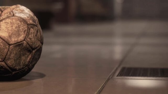 A worn soccer ball sits on a smooth floor close to a drain. The scene shows signs of indoor play, with little light and a quiet environment.