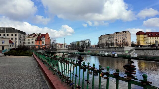 Video of a green railing covered with love padlocks along a river in an urban area, featuring historic city buildings, a bridge, and a blue sky with white clouds on a sunny day.