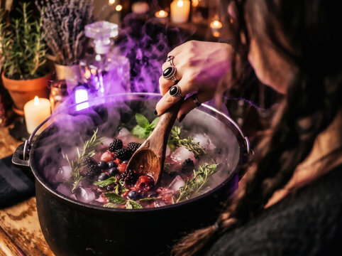Witch stirring a bubbling purple magic potion with blackberries blueberries herbs and dry ice smoke in a black cauldron on a rustic wooden table for Halloween