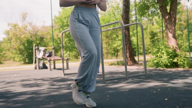 Close up of young woman performing high knee exercise in outdoor park setting. Woman wears gray sweatpants and white sneakers with green trees visible behind her. scene is bright daylight