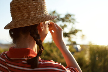 Naklejka premium Young woman in a straw hat with braided hair looks through binoculars at the sunny meadow. An authentic lifestyle scene captures a nature adventure and exploration.