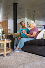 Naklejka premium Diverse female pair sitting on dark gray couch, senior holding smartphone, companion leaning in