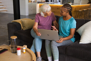 Naklejka premium Senior woman and African American woman sitting on sofa at home using laptop with pill bottles