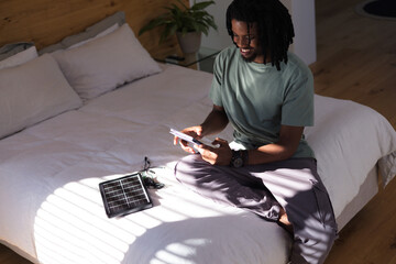 African American man sitting cross-legged on bed at right holding phone and solar panel, copy space © WavebreakMediaMicro