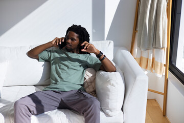 Fototapeta premium African American man reclining on white sofa at home, wearing sage-green tee, holding smartphone