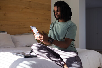 Man sitting on bed cross-legged wearing sage shirt holding lit tablet, charging cable connecting © WavebreakMediaMicro