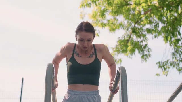 Close up of woman performing step up exercise on metal stairs outdoors with green foliage and clear sky in background. Woman wears dark sports bra and light gray shorts holding railings