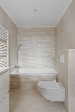 Minimalist modern bathroom interior with bathtub, wall mounted shower, beige textured tile, wall hung toilet, bright neutral finishes, clean residential layout, and refined spa inspired comfort