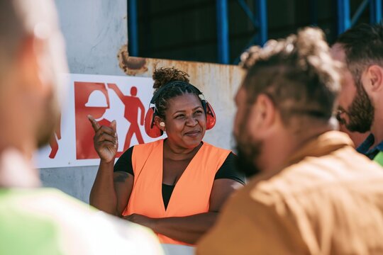 Black woman in high-visibility vest briefing a crew by a safety panel, natural industrial setting with cool for EHS training and workplace safety