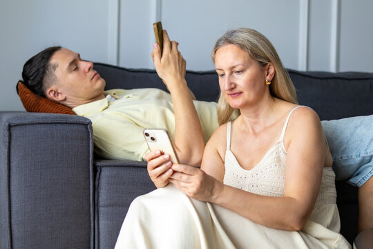 Middle-aged woman sits beside man as both look at their own phones together on couch. Concept of shared screen time and parallel phone use during couple relaxation at home.