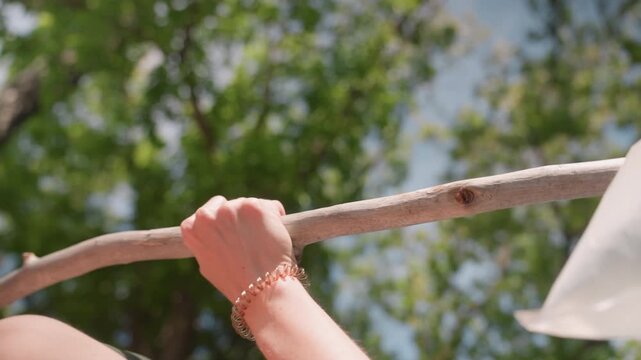 Close up of person hands gripping and moving along tree branch with sunlight filtering through green leaves. subject wears bracelets on their wrists one metallic chain bracelet visible
