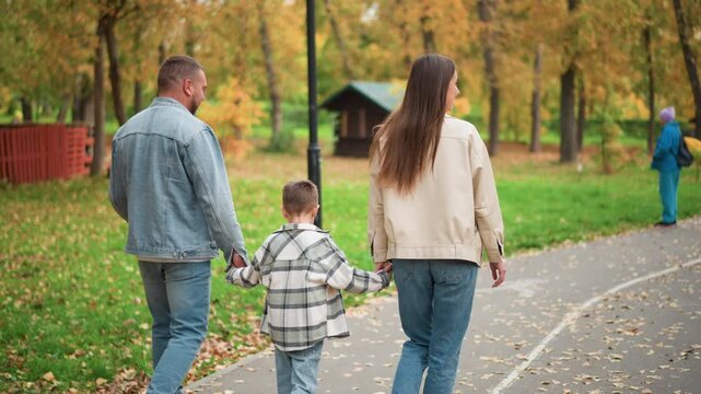 Family of three walking together in park holding hands as they stroll along path surrounded by autumn foliage. scene features green grass with fallen leaves trees displaying fall colors