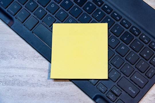 Top view of square yellow sticky note placed on a black computer keyboard on wooden desk background. Copy space for text or mockup.