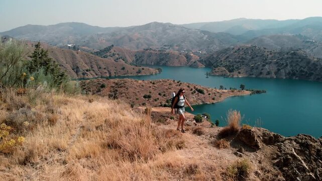Woman hiking steadily along a rugged mountain ridge, embracing solitude and freedom on a serene turquoise reservoir under a clear sky, discovering self-reliance and personal growth through outdoor adv