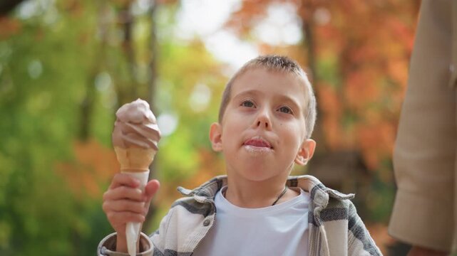 Young boy enjoying ice cream cone outdoors in park setting holding treat and taking bites while surrounded by autumn foliage. Boy wears plaid jacket over white shirt with soft lighting