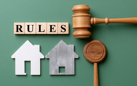 Wooden judge gavel next to letter blocks spelling rules and small house models on green background symbolizing real estate law and property code
