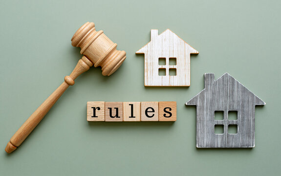 Wooden judge gavel next to letter blocks spelling rules and small house models on green background symbolizing real estate law and property code
